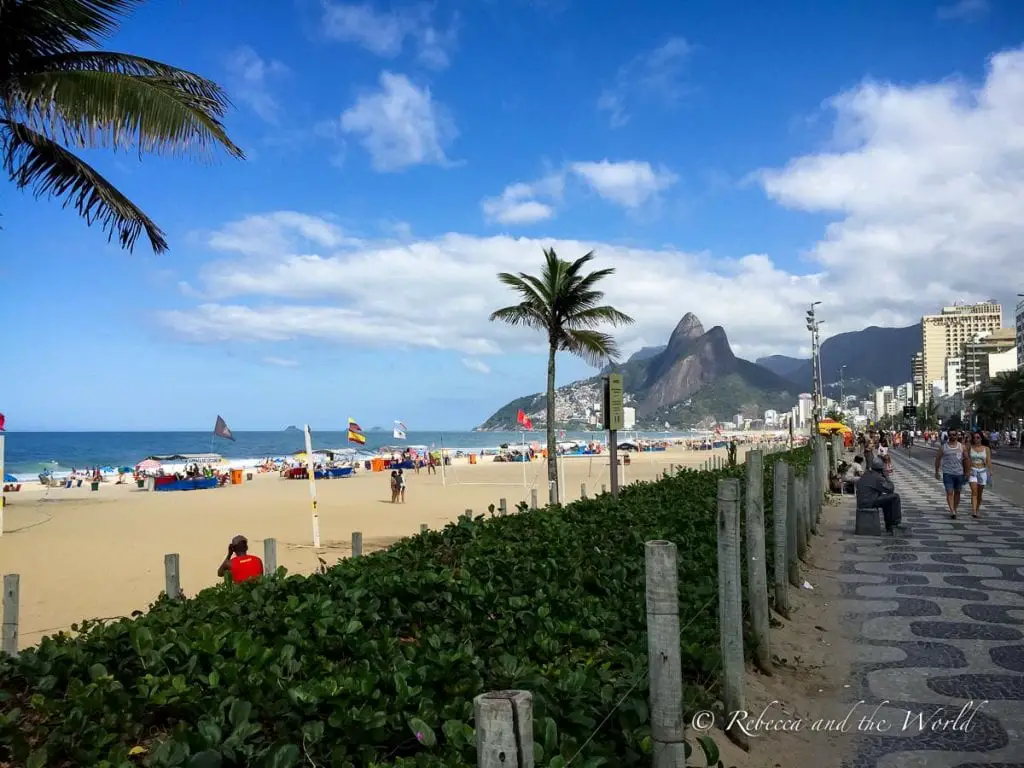 A sunny beach scene with a blue sky, palm trees, and people enjoying the sand and surf. A mountain looms in the background. Rio de Janeiro is one the best places to visit in Brazil, and there's plenty to do in 3 days.