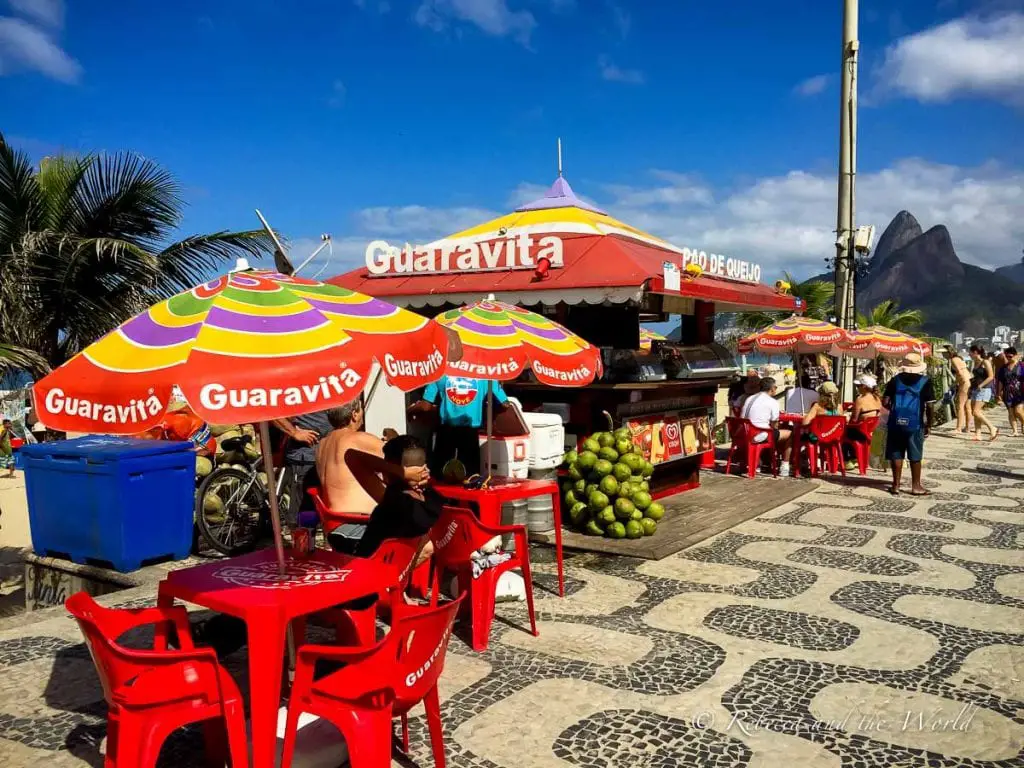 A colorful beachfront scene with red chairs and umbrellas, labeled "Guaravita." A street vendor's stand offers snacks under a bright blue sky, with a mountain visible in the distance. Copacabana is one of Rio de Janeiro's most famous beaches - and a Rio must-visit.