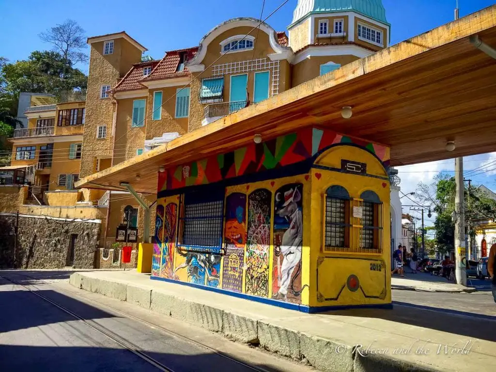 A colorful tram station painted with vibrant murals, with a yellow tram painted on its façade. The background features a multi-storied building with a tiled roof. Take a bonde, a historic tram, up to Santa Teresa to wander through this lovely neighbourhood in Rio de Janeiro.