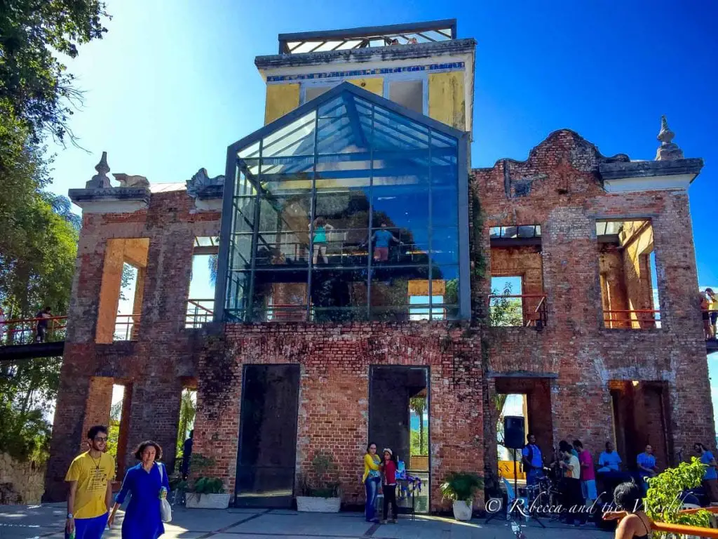 A ruined brick building with no roof, partially restored with a modern glass structure. People are walking around, exploring the site. Parque das Ruinas in Santa Teresa, Rio de Janeiro, is home to a beautiful old mansion.