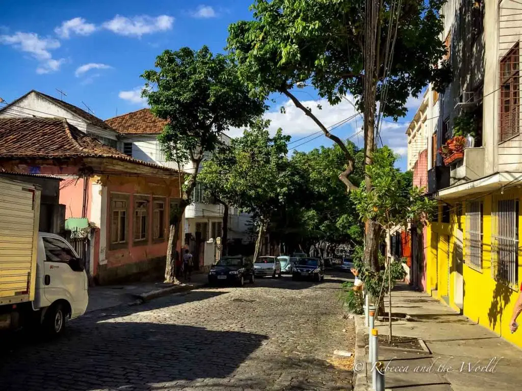 A quiet street scene with old, colorful houses on either side and trees providing shade. The road is cobbled, and a few cars and people are visible. Put a visit to Santa Teresa on your Rio de Janeiro itinerary - it's a lovely bohemian neighbourhood.
