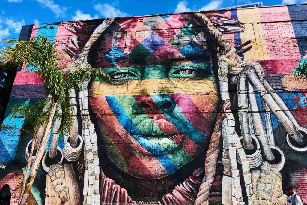 A large, intricate mural of a woman's face painted in a multitude of colors on a building, flanked by palm trees. The artwork has a vibrant and detailed appearance. Mural Etnias is a 30,000-square-foot mural in Rio de Janeiro that represents the cultural diversity of the Olympic Games.