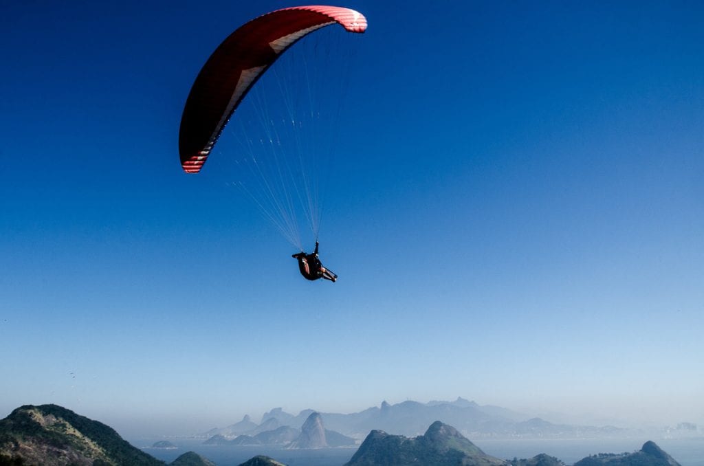 A paraglider soaring in a clear blue sky, with distant mountains and a coastline beneath. Paragliding is a popular thing to do in Rio de Janeiro.