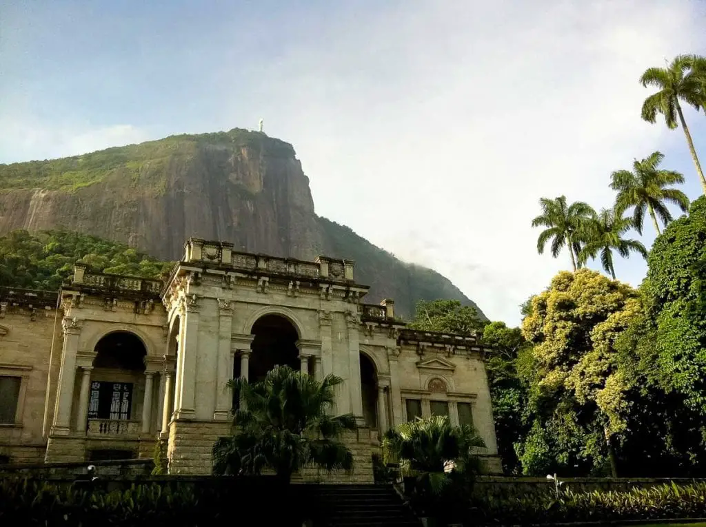An old classical building with pillars in the foreground, dense greenery surrounding it, and a mountain with the Christ the Redeemer statue at the peak in the background under a bright sky. Parque Lage is a photogenic public park in Rio de Janeiro, with an old mansion.