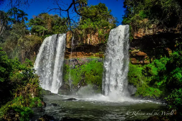 Two powerful waterfalls at Iguazu Falls, surrounded by cliffs and greenery, with water forcefully dropping into a pool below creating a cloud of spray.