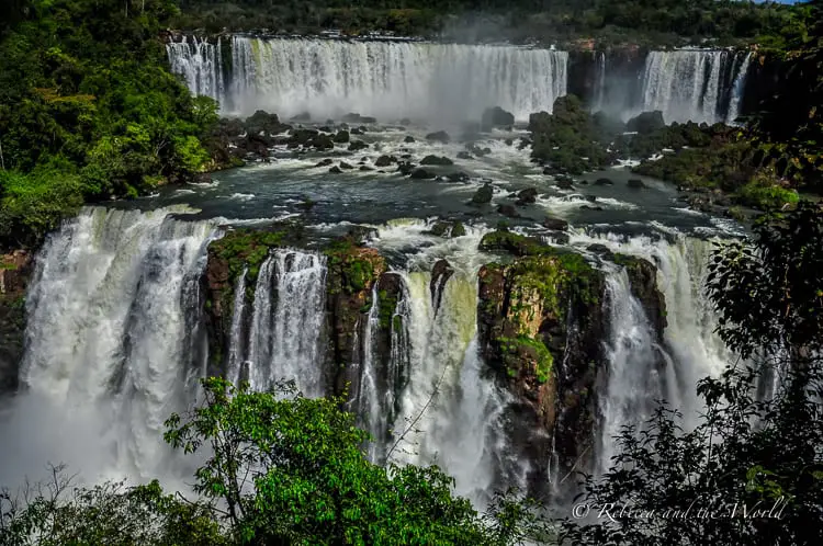 A closer view of a section of Iguazu Falls showing multiple cascades of water plunging over rugged cliffs surrounded by dense forest, with mist rising from the base of the falls.