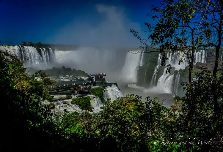 A view of Iguazu Falls in Brazil with a vast amount of water cascading down multiple levels of cliffs. The falls are partially shrouded in mist, with a viewing platform visible where people can be seen observing the natural spectacle.