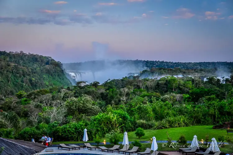 A tranquil scene captured at dusk showing the distant Iguazu Falls enveloped in mist, viewed from luxury hotel Gran Melia Iguazu in Argentina with lounge chairs in the foreground and a dense tropical forest surrounding the area.