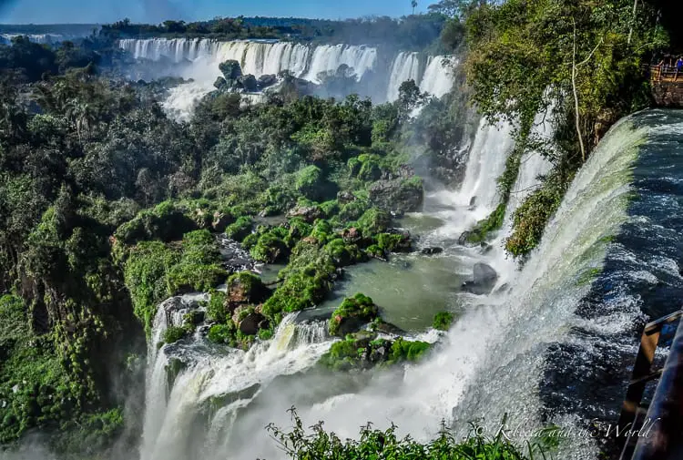 An angled perspective of Iguazu Falls in Brazil where the water powerfully rushes over a precipice, surrounded by lush greenery. A viewing platform on the right is filled with spectators taking in the scene.