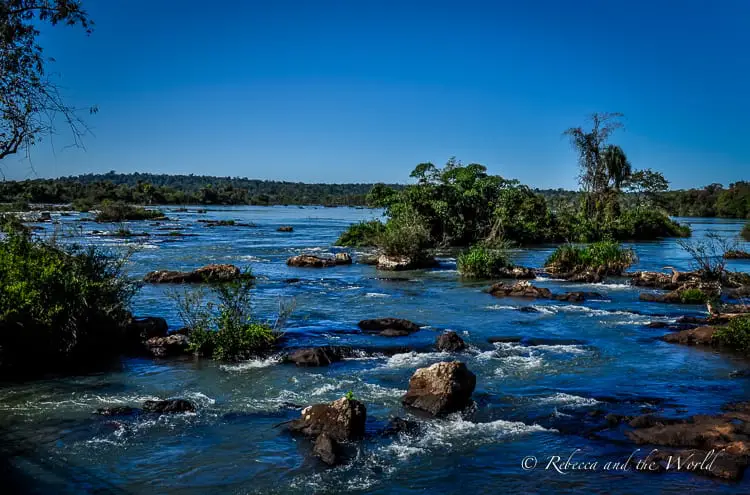 The upstream view of Iguazu River dotted with small islands covered in thick vegetation, under a clear blue sky, just before the water plunges over the falls. Should you visit Iguazu Falls Argentina or Brazil side? I say both!