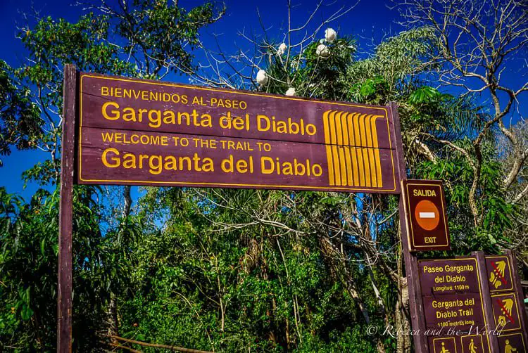 A wooden signboard for "Garganta del Diablo" or "Devil's Throat," a section of Iguazu Falls, with directional information and a welcome message in Spanish and English. This guide includes the latest information about visiting Iguazu Falls from both the Argentina and Brazil sides.