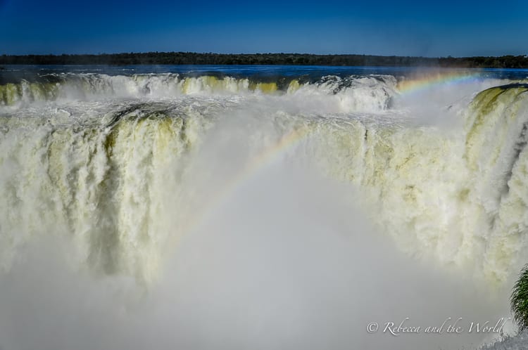 A dramatic close-up of the crest of Iguazu Falls, where a massive volume of water plummets into a misty abyss. A rainbow is visible through the spray at the bottom of the falls.