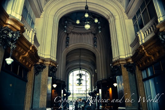 Interior shot of the grand entry hallway in Palacio Barolo. The space is tall with arched ceilings, ornate columns, and hanging chandeliers. The hallway leads to a backlit glass door at the far end.