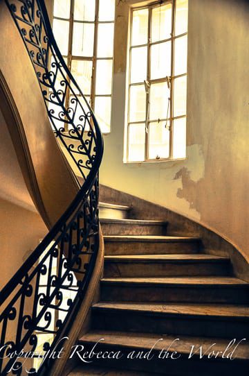 A staircase inside Palacio Barolo with worn marble steps and a delicate wrought-iron railing. A large window with natural light illuminates the stairwell, highlighting the patina on the walls and the staircase's graceful curve.