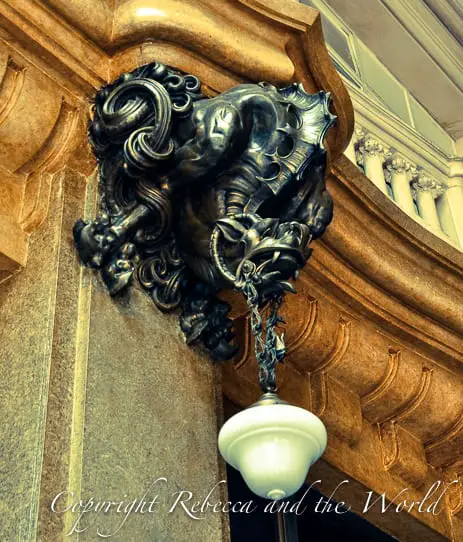 Close-up of a sculpted bronze bracket holding a white glass lamp. The bracket has a figure resembling a mythological creature with detailed swirls and flourishes, set against a marble wall. This is one of the interesting and intricate details inside the Palacio Barolo building in Buenos Aires.