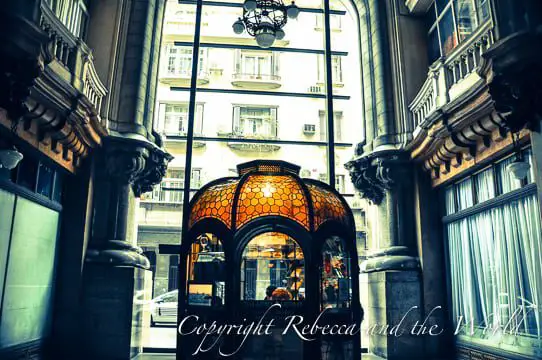 View inside Palacio Barolo featuring a domed information booth with a striking stained glass top in warm orange tones. The booth is framed by grand columns and a large glass window that allows natural light to filter in.