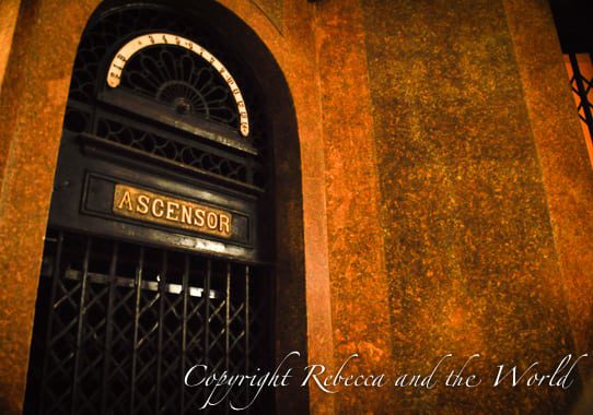 The ornate black metal gate of an elevator, labeled 'ASCENSOR' in golden letters, with a half-circle fanlight above it. The elevator is set in a marble wall with a warm glow from the ambient lighting.