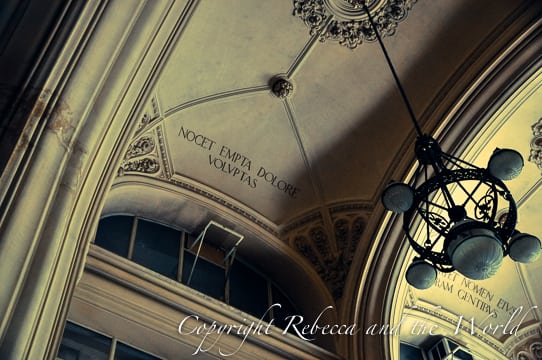 Interior shot capturing a ceiling with a chandelier and inscriptions around an arch. The Latin phrases and decorative elements add to the historical ambiance of the space.