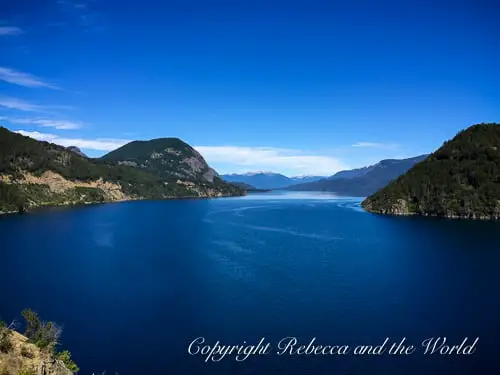A serene lake landscape in Bariloche, Argentina, along the Ruta de los Siete Lagos, with deep blue water, nestled between forested hills under a bright blue sky.