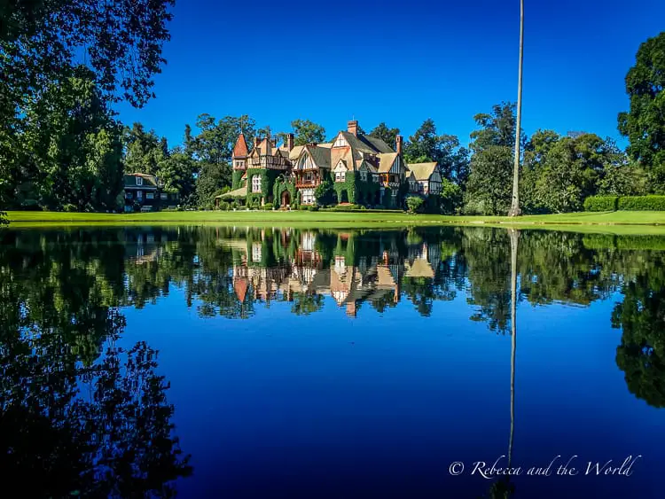 A picturesque Tudor-style mansion reflected in a tranquil pond, surrounded by a manicured lawn and trees under a clear blue sky. The property is Estancia Villa Maria, the author of this article's favourite estancia near Buenos Aires.