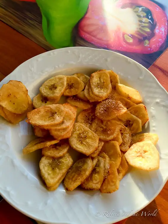 A white plate filled with fried plantain chips (or tostones), seasoned lightly with salt, set against a colorful background of a picture of a tomato and green pepper.