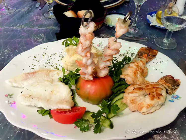 A platter of assorted seafood including grilled fish fillets, skewered shrimp, and lobster tail, garnished with fresh parsley, tomato slices, and cucumber slices.