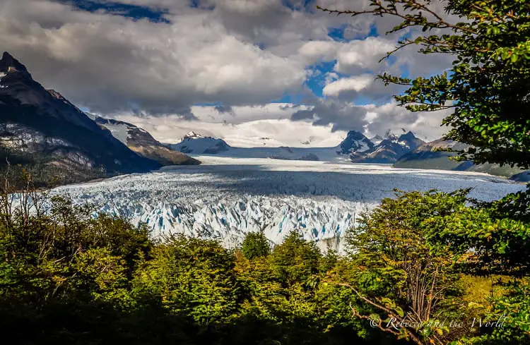 The vast Perito Moreno glacier framed by forested slopes under a cloudy sky. The ice field extends into the distance, flanked by rugged mountains. Trekking Perito Moreno Glacier is one of the best things to do in Argentina.