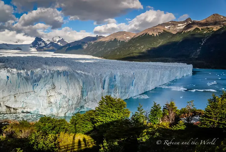 A panoramic view of the massive Perito Moreno glacier flowing into a lake. The glacier's surface is a vivid blue-white, and it is surrounded by dark green foliage and distant mountains under a partly cloudy sky.