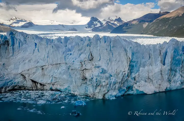 A close-up of the icy face of the Perito Moreno Glacier. The glacier’s intricate textures and shades of blue stand out against the surrounding water and snow-capped mountains in the distance. One of the most incredible things to do in Argentina is go trekking on Perito Moreno Glacier.