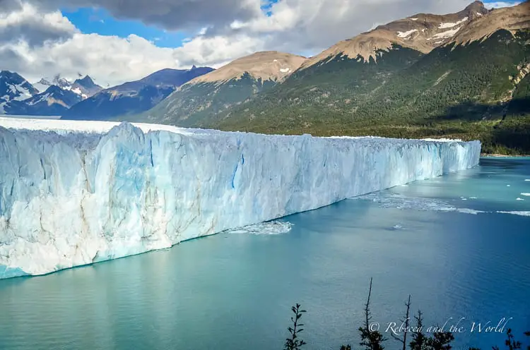 The side view of the towering Perito Moreno glacier wall meeting the blue waters of a lake. The ice structure is massive, with a backdrop of mountainous terrain. This glacier is one of the most beautiful places to visit in Argentina - and trekking Perito Moreno Glacier is even more incredible!