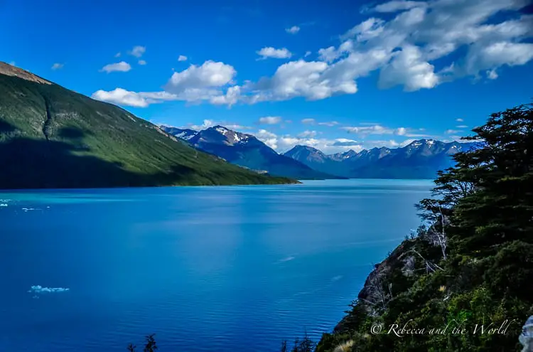 A serene lake with a scattering of icebergs, bordered by greenery and mountains stretching under a partly cloudy sky.