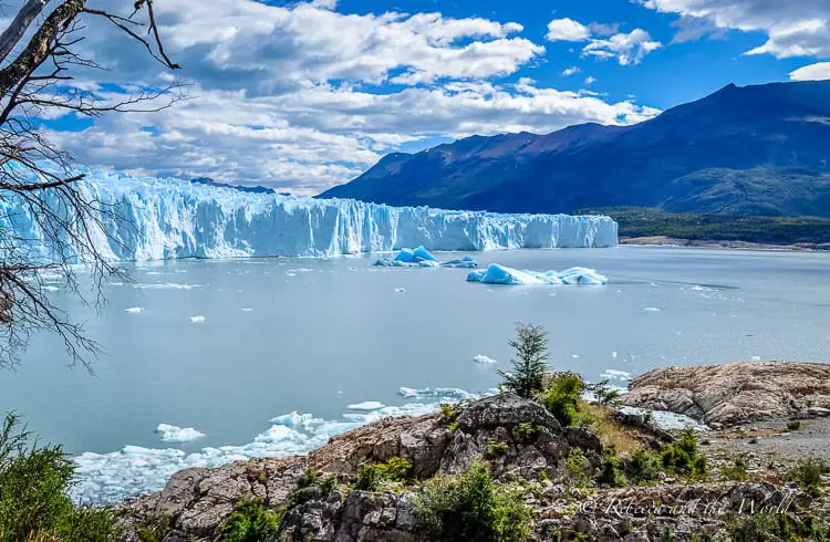 A view of a glacier front from across a lake, with chunks of ice floating on the water, flanked by rugged hills under a blue sky.