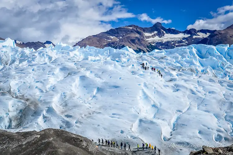 A group of hikers on an ice field, dwarfed by the glacier's vast, undulating surface. Snow-capped mountains rise in the background. Trekking Perito Moreno Glacier is one of the most incredible things to do in Argentina.