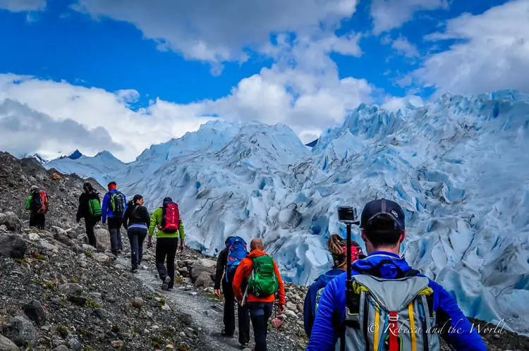 Hikers trekking on a rocky path towards Perito Moreno glacier in Argentina, with the ice's rugged surface visible ahead. They are equipped with backpacks and colorful attire.