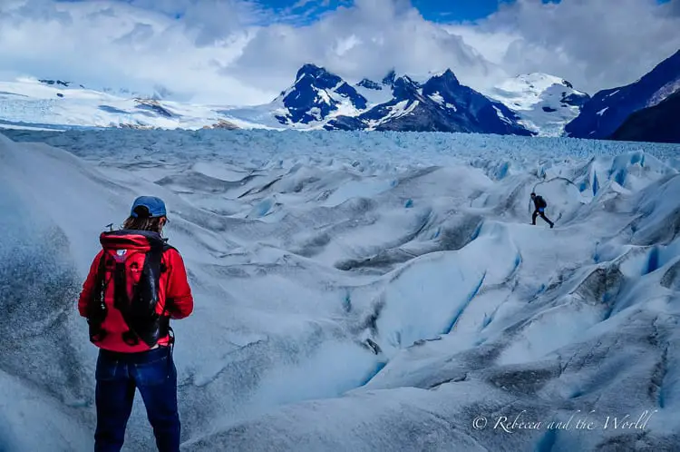 A lone hiker in red looks out over a crevassed glacier expanse. In the distance, other hikers can be seen exploring the ice. This is the incredible Perito Moreno Glacier in Argentina.