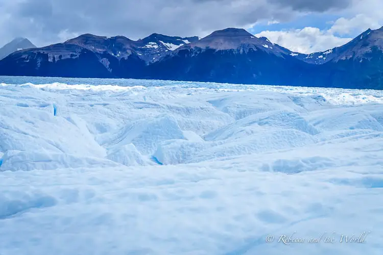 A vast, textured surface of a glacier with deep crevasses and ridges, set against a backdrop of dark mountains and a cloudy sky. This is the view of Perito Moreno Glacier in Argentina.