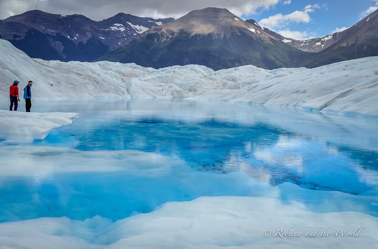 Two people stand beside a deep, vividly blue pool of water on a glacier, surrounded by the white and blue of the ice field with mountains beyond.