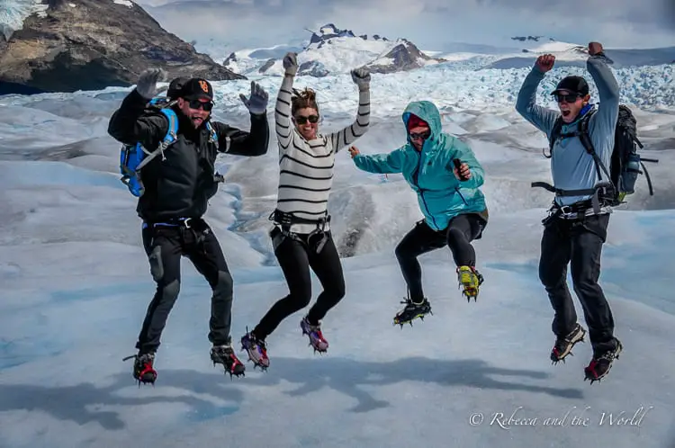 Four people - including the author of this article - in warm clothing and crampons joyfully jumping on Perito Moreno glacier with snow-covered peaks in the background.