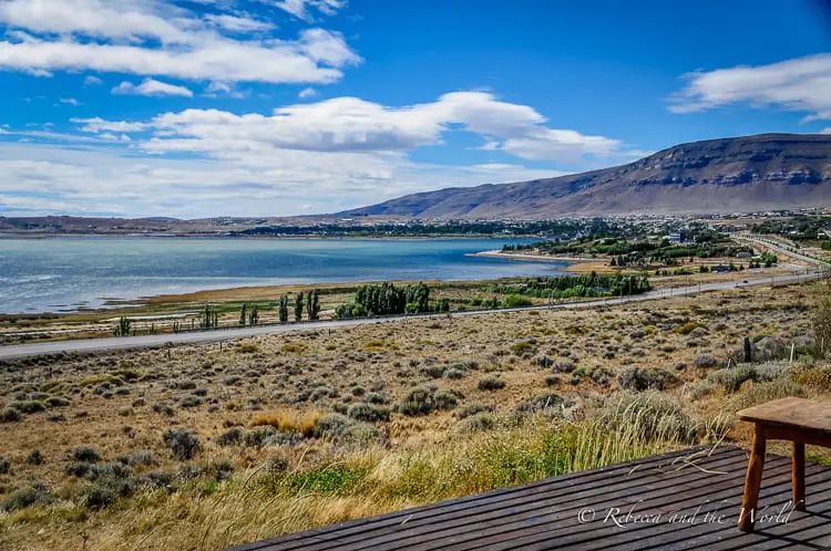 A landscape view featuring a tranquil lake, bordered by a small town and roads, set against rolling hills and a blue sky with scattered clouds. This is the view from the author's hotel in El Calafate, Argentina.