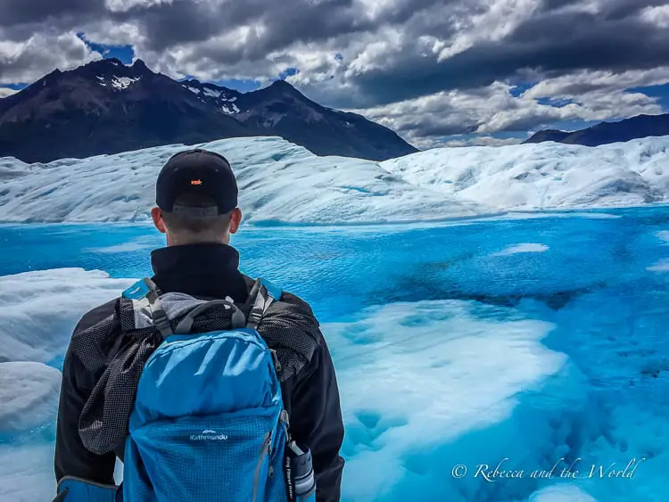 A person - the author's husband - with a backpack standing before a deep blue glacial pool, with the expansive white glacier and dark mountains under a stormy sky. This is what you'll see while trekking Perito Moreno Glacier in Argentina.