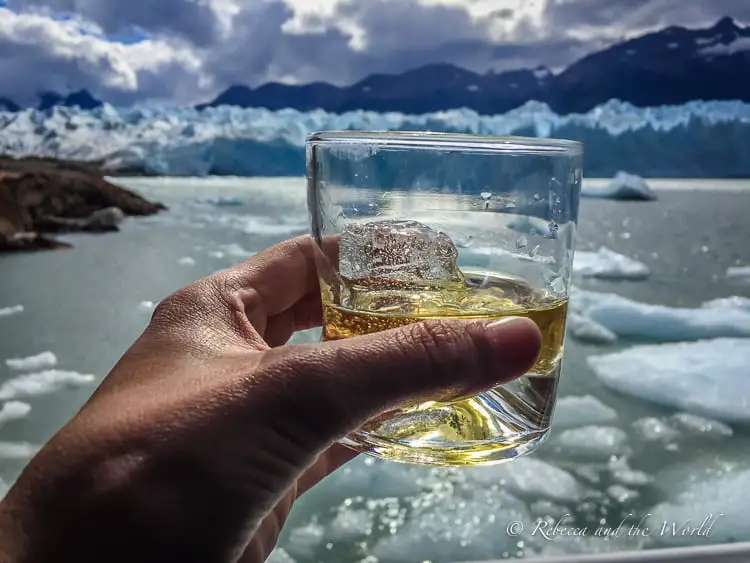 A hand - belonging to the author of this article - holds a glass with amber liquid and ice, with a blurred glacier and mountainscape in the background. A glass of whiskey is one of the rewards at the end of trekking Perito Moreno Glacier in Argentina.