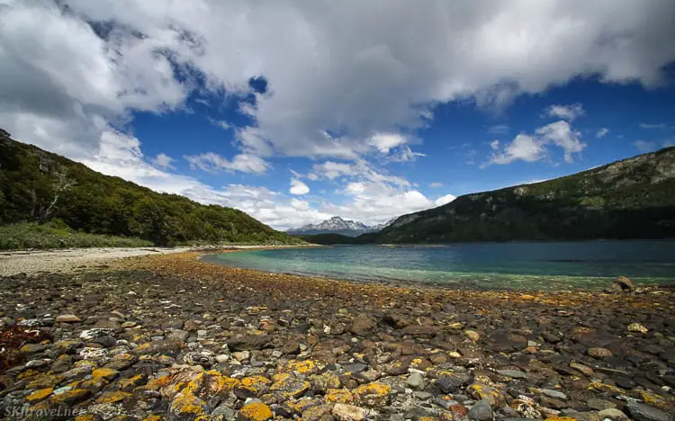 A pebbled beach leading to clear, shallow waters with mountains in the distance under a partly cloudy sky. This is Tierra del Fuego National Park, one of the best places to visit in Ushuaia, Argentina.