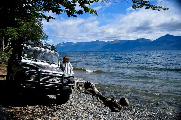 A rugged off-road vehicle parked by a lakeside with a woman standing by the shore, looking out at the water and distant mountains. There are so many beautiful places to visit in Ushuaia, including a lot of outdoors destinations.