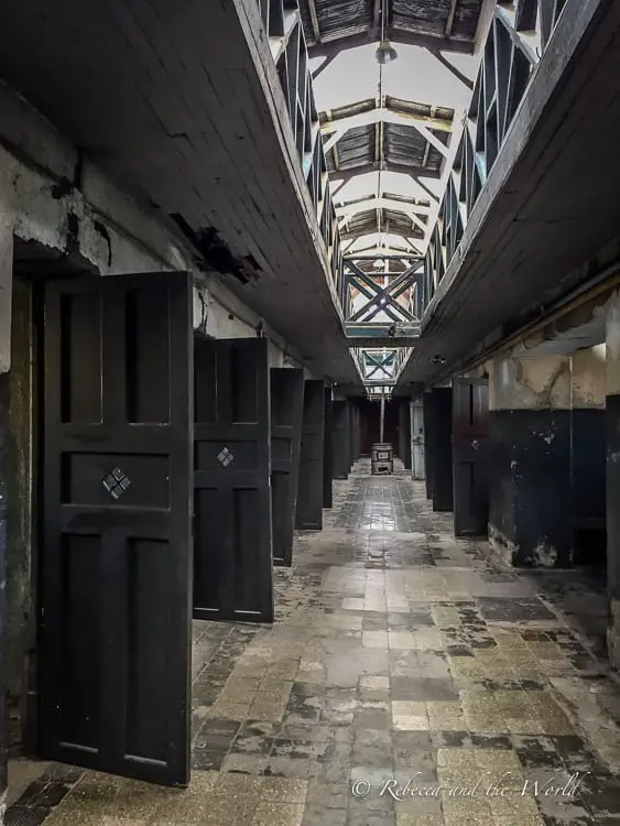 An old wooden corridor with rows of closed doors on either side, dimly lit from above. This is the Prison Museum in Ushuaia, an important historical tourist attraction.