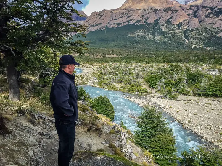 A tranquil scene of a man - the author's husband - in black hiking gear standing on the edge of a cliff overlooking a river with a dense forest leading up to rugged mountains in the distance.