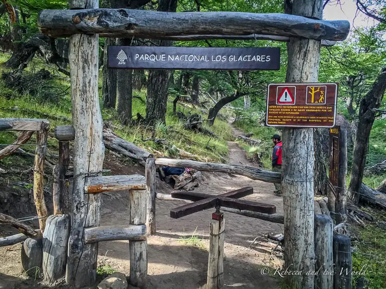 The entrance to Parque Nacional Los Glaciares, marked by a rustic wooden gate with a sign overhead. A cautionary sign to the right advises hikers about trail safety and conduct.