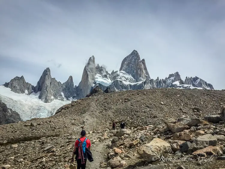 Hikers on a rocky path with the sharp, towering peaks of Mount Fitz Roy in the distance. The mountain's dramatic profile and glaciers are a stark contrast against the barren landscape in the foreground.