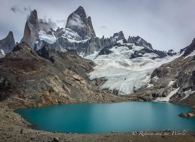 The serene blue waters of a glacial lake in the foreground, with the imposing Mount Fitz Roy and its surrounding glaciers towering in the background under a cloudy sky.