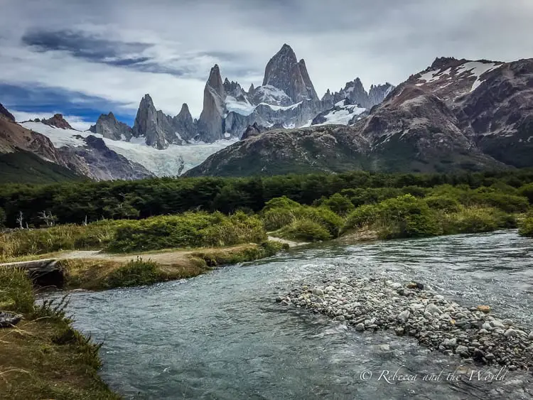 Scenic view of a clear river winding through a lush valley with the majestic, jagged peaks of Mount Fitz Roy in the distance. Overcast skies loom above the dramatic landscape, typical of El Chaltén's rugged terrain.