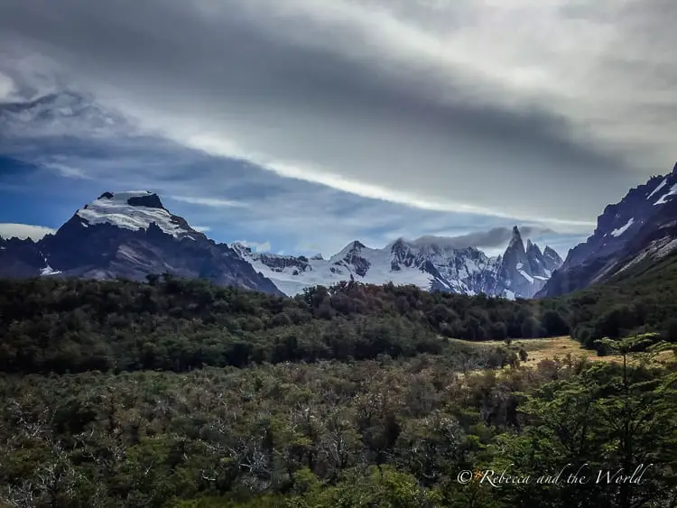 A breathtaking vista of a forest opening up to reveal the imposing, snow-capped peaks of the El Chalten, with clouds casting shadows over the mountains, creating a dramatic and beautiful natural scene.